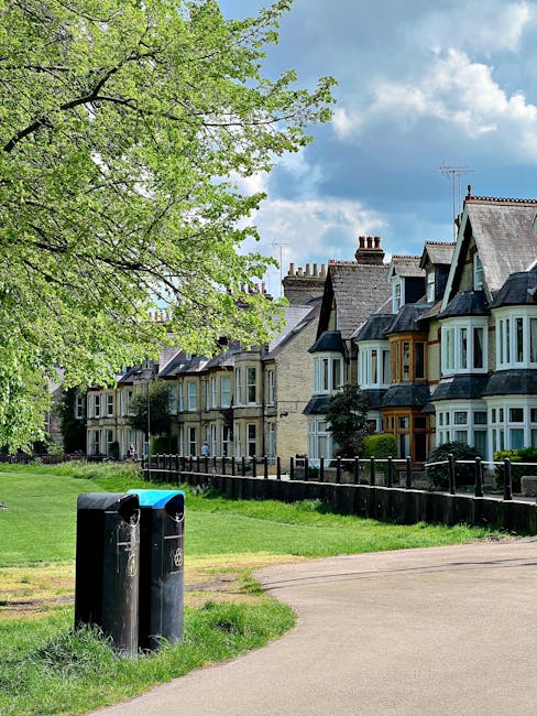 A residential street scene featuring a row of traditional terraced houses with bay windows, tiled roofs, and chimneys, set behind a well-maintained garden with trimmed hedges and lush green grass. In 
